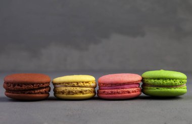 Koyu gri bir zeminde arka arkaya çok renkli badem kurabiyeleri. Gri beton arka planda makaronlar. Pastalar üst üste. Metin için boşluk, kopyala boşluk.