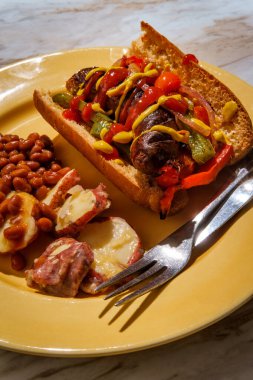 Sausage onions and peppers hoagie sandwich with side of baked beans and German potato salad clipart