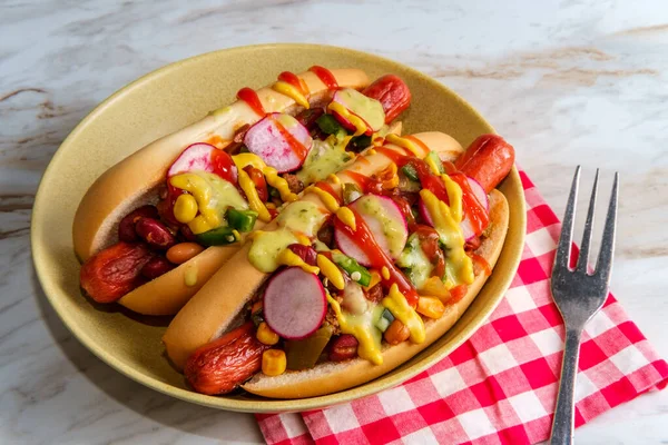 Fancy chili dogs topped with radishes mustard ketchup and green sauce on gluten-free buns