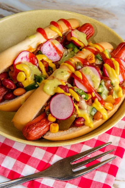 Fancy chili dogs topped with radishes mustard ketchup and green sauce on gluten-free buns