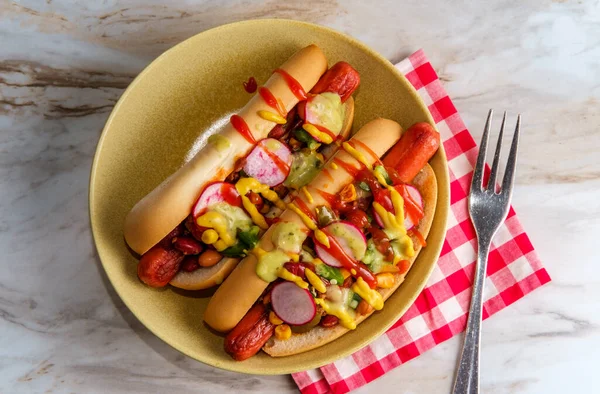 Fancy chili dogs topped with radishes mustard ketchup and green sauce on gluten-free buns