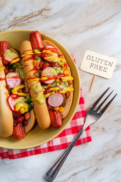 Fancy chili dogs topped with radishes mustard ketchup and green sauce on gluten-free buns