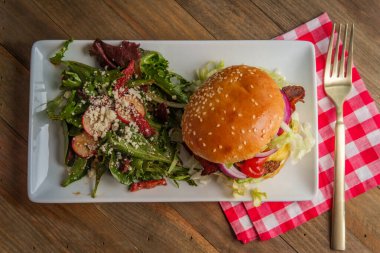 Yanında Yunan salatasıyla servis edilen gevrek domuz pastırmalı Angus bifteği çizburgeri.