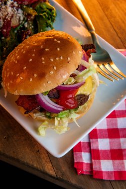 Yanında Yunan salatasıyla servis edilen gevrek domuz pastırmalı Angus bifteği çizburgeri.