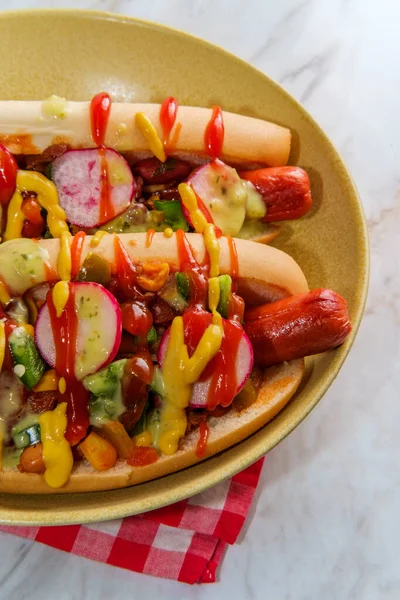 Fancy chili dogs topped with radishes mustard ketchup and green sauce on gluten-free buns
