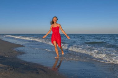 Genç mutlu kadın sahilde koşuyor ve hayatın tadını çıkarıyor. Kırmızı elbiseli gülümseyen kız deniz kıyısında koşuyor.