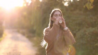 Elinde kahve fincanıyla sonbahar parkında yürüyen güzel bir kadın. Dışarıda gün batımında yürümek, sıcak güneş ışığı, sıcak sonbahar havası..