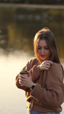 Genç bir kadın gün batımında nehrin kenarında sıcak kahveyle ısınıyor. Sonbahar sabahı serinliği, rahat ruh hali, rahatlama, doğa ve sakinlik..