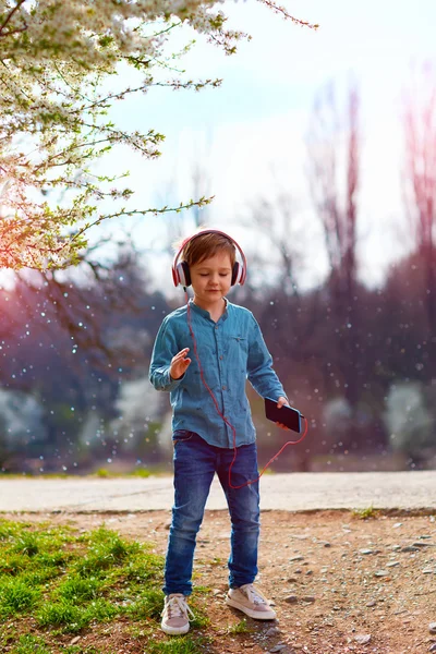 Χαριτωμένο παιδί αγόρι με ακουστικά ακούει τη μουσική και το χορό σε ανθισμένο πάρκο — Φωτογραφία Αρχείου