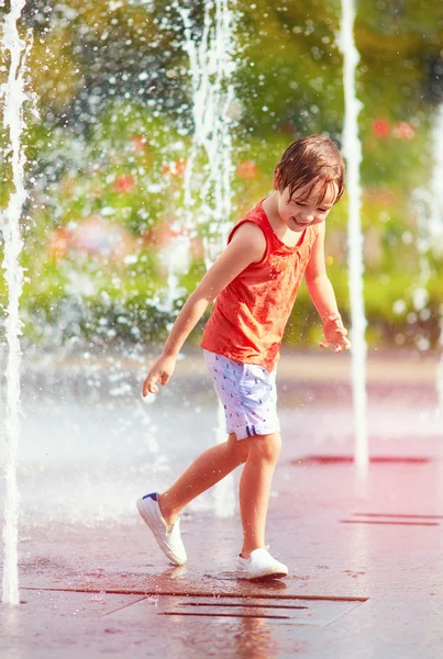 Glada pojke med kul mellan vattenstrålarna, i fontänen. Sommar i staden — Stockfoto