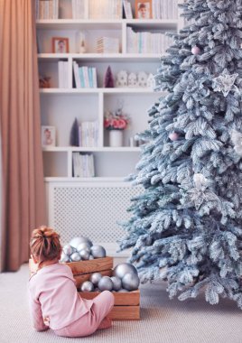 Küçük bir kız bebek evde, halının üzerinde bir Noel ağacının önünde Noel süsleriyle oynuyor.