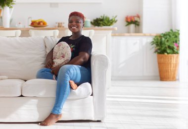 Evdeki güneşli oturma odasındaki kanepede oturan neşeli genç kadın.