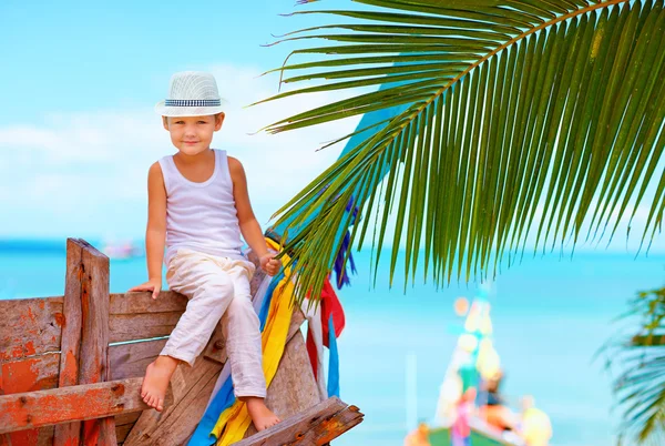 Χαριτωμένο μοντέρνα αγόρι που θέτουν σε παλιά βάρκα στο τροπική παραλία — Φωτογραφία Αρχείου