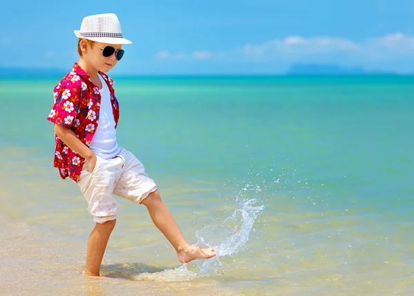 Happy fashionable kid boy walking in surf on tropical beach — Stock Photo, Image