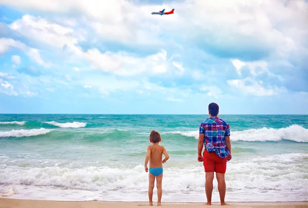 Отец и сын стоят на пляже в штормовую погоду, наблюдая, как летит самолет — стоковое фото