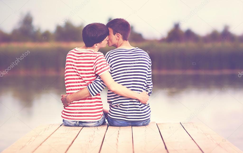 male couple, kissing, while sitting on wooden pier at riverbank