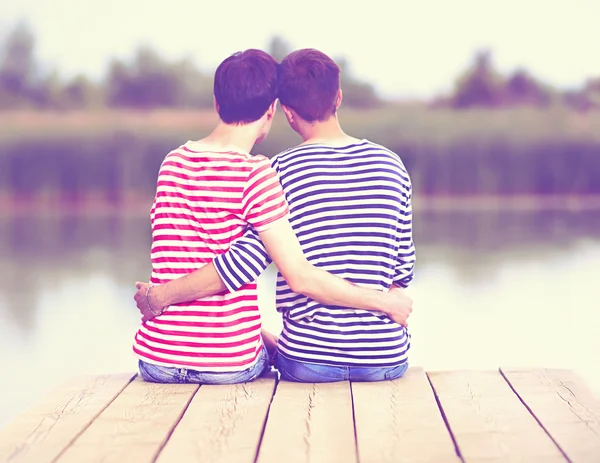 Male couple in love, sitting on wooden pier at riverbank — 图库照片