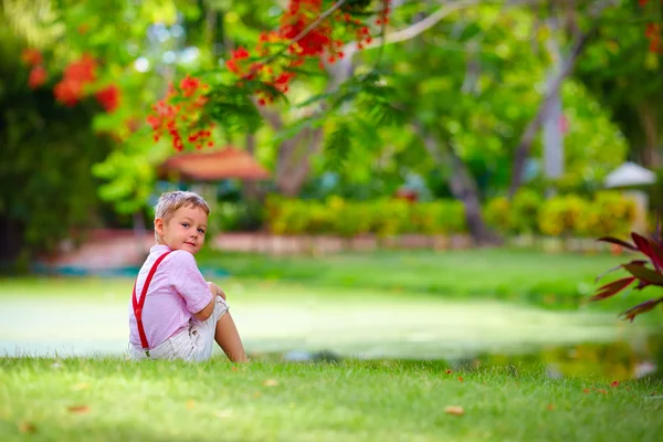 Mignon enfant assis sur la pelouse verte près du lac tropical — Photo