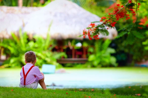 Mignon enfant assis sur la pelouse verte près du lac tropical — Photo