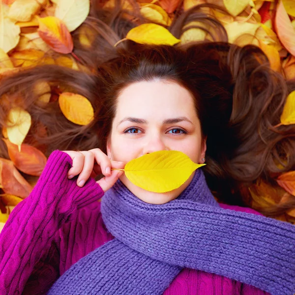 Young girl lying among autumn leaves, hiding lips behind a leaf — Stock Photo, Image