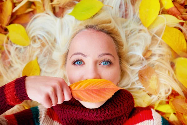 Young girl lying among autumn leaves, hiding lips behind a leaf — Stock Photo, Image