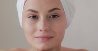 Kahverengi gözlü çekici kadın ellerine yaslanırken doğrudan kameraya bakıyor. Kafasında beyaz banyo havlusu olan genç bir kadın. Cilt bakımı kavramı.