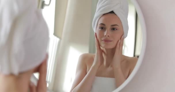 Mulher bonita envolto em toalha de banho vendo sua pele rosto saudável e brilhante após o chuveiro da manhã. Reflexão no espelho. Procedimentos de beleza. — Vídeo de Stock