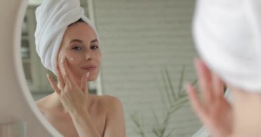 Banyoda duran ve yüzüne krem süren beyaz elbiseli mutlu kadın. Çıplak omuzlu genç bir kadın cilt bakımı için Kore kozmetik ürünleri kullanıyor..