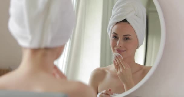 Hermosa hembra envuelta en toalla blanca hidratando su piel durante la rutina matutina. Mujer joven con belleza natural utilizando almohadilla de algodón en el baño moderno. — Vídeo de stock
