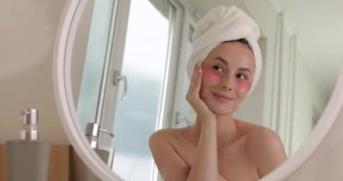 Yüzünde kozmetik bantlarla banyo havlusu olan mutlu ve çekici bir kadın aynaya bakıyor ve evde sabahları eğleniyor. Sağlık ve vücut bakımı kavramı.