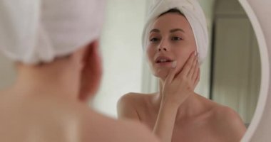 Kafasında banyo havlusu olan çekici genç bayan aynaya bakarken temiz yüz derisinden memnun görünüyor. Güzellik ve sabah rutini.