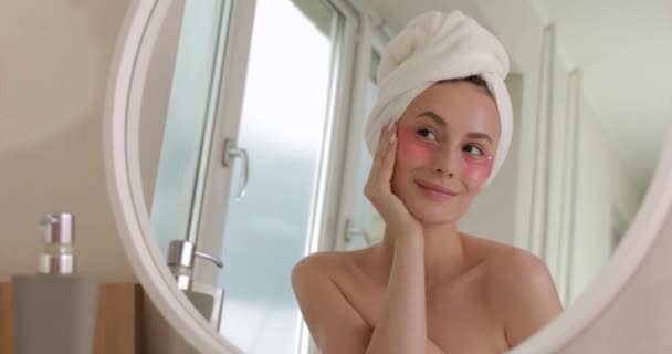 O femeie atractivă fericită în prosop de baie cu plasturi cosmetici sub ochi uitându-se la oglindă și bucurându-se de ora de dimineață acasă. Conceptul de sănătate și îngrijire corporală . — Videoclip de stoc