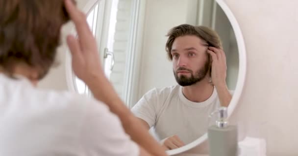 Glad skäggig man i vit skjorta står på ljust badrum, ser och spegel och poppar finnar. Hudvårdsförfaranden under morgonen. — Stockvideo