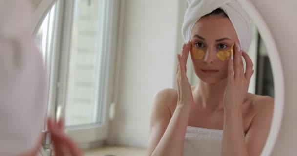 Ładna młoda dama z białym ręcznikiem na głowie przy użyciu koreańskich plastrów na oczy po porannym prysznicu. Pojęcie pielęgnacji skóry i kobiet naturalne piękno — Wideo stockowe