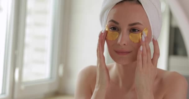 Adorabilă brunetă tânără în prosop de baie folosind plasturi aurii pentru ridicarea pielii sub ochi. Reflecție în oglindă. Proceduri de frumusețe de dimineață . — Videoclip de stoc