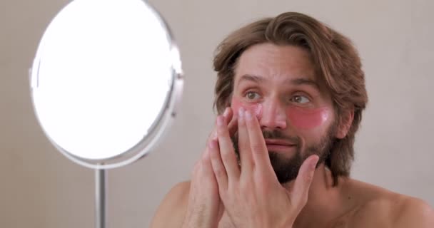 Bonito barbudo cara com ombro nu usando manchas cosméticas sob os olhos enquanto olha para o espelho. Conceito de saúde masculina e anti envelhecimento. — Vídeo de Stock