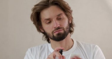 Beyaz tişörtlü sakallı genç adam pamuk ped ve profesyonel makyaj malzemesi kullanıyor. Yakışıklı bir adam evde cildiyle ilgileniyor..