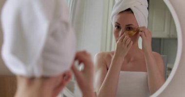 Kafasında banyo havlusu olan güzel genç bir kadın aynaya bakıyor ve altın göz bandı kullanıyor. Siyah halkaları göz altında küçültmek için güzellik prosedürleri yapan mutlu bayan..