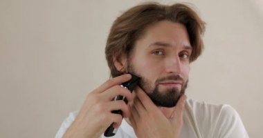 Beyaz tişört giymiş, sakalını elektrikli traş makinesiyle tıraş eden çekici bir gencin portresi. Bakım, temizlik ve güzellik anlayışı..