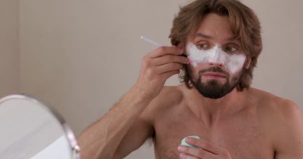 Pozytywny brodaty mężczyzna za pomocą szczotki kosmetycznej do stosowania białej maski gliny na twarz. Pojęcie ludzi, pielęgnacja i pielęgnacja skóry. — Wideo stockowe