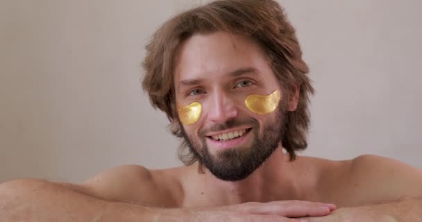 Felice uomo barbuto con le spalle nude che indossa toppe sotto gli occhi. Giovane ragazzo che utilizza cosmetici coreani per sollevare la pelle e ridurre le rughe. — Video Stock