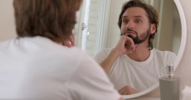 Счастливый молодой человек в белой рубашке проводит утро в ванной и смотрит в зеркало. Бородатый мужчина выглядит удовлетворенным состоянием кожи лица. — стоковое видео
