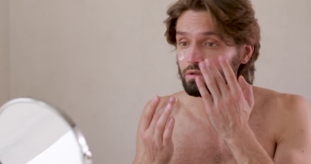 Hombre joven sin camisa mirando al espejo pequeño y aplicando crema facial. Hombre barbudo usando cosméticos profesionales para hidratar la piel. — Vídeos de Stock