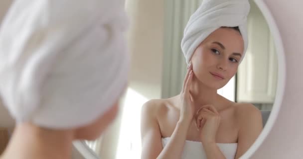Szczęśliwa młoda kobieta owinięta w ręcznik, patrząca w lustro i ciesząca się świeżą skórą po porannym prysznicu. Rytuały piękności w domu. — Wideo stockowe