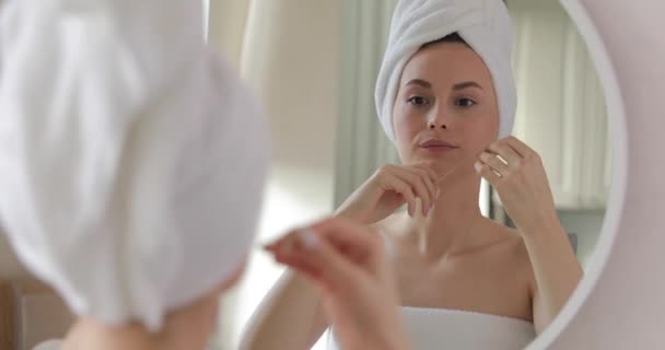 Mujer joven y feliz envuelta en toalla blanca de pie en el baño luminoso, mirando al espejo y limpiando los dientes con hilo dental. Concepto de higiene bucal. — Vídeos de Stock
