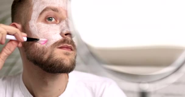 Młody człowiek z brodą nakładający maskę nawilżającą na twarz pędzlem po prysznicu. Szczęśliwy mężczyzna w białej koszulce robi zabiegi pielęgnacji skóry w domu. — Wideo stockowe
