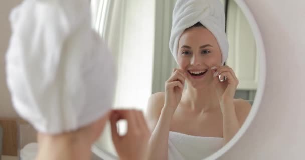 Jovem atraente com toalha de banho na cabeça e no corpo usando fio dental enquanto passa o tempo da manhã no banheiro. Reflexão no espelho. Higiene oral em casa. — Vídeo de Stock