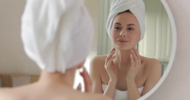 Приваблива молода жінка з голими плечима, яка дивиться на шкіру обличчя, стоячи у ванній кімнаті. Відбиття у дзеркалі. Концепція догляду за шкірою . — стокове відео