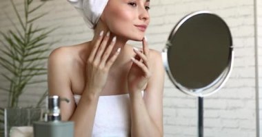 Duştan sonra beyaz havluya sarılmış güzel bir kadın aynaya bakıyor ve yüzüne dokunuyor. Doğal güzelliği olan mutlu kadın cilt hastalığından zevk alıyor..