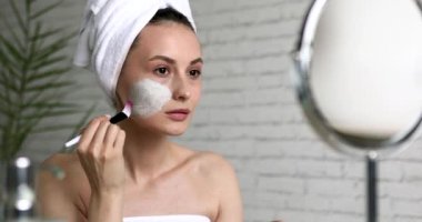 Banyo havlusu giymiş rahat genç bir kadın aynaya bakarken yüzüne fırçayla maske takıyor. Günlük güzellik rutini. Evde yüz bakımı..